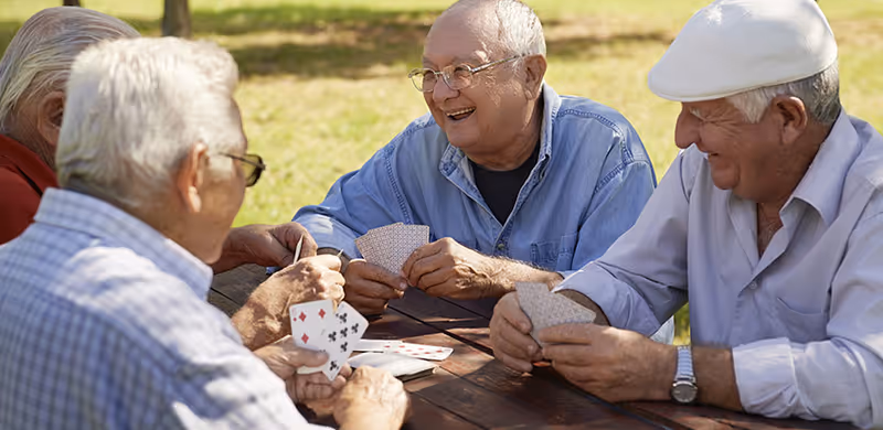 A group of elderly men sitting around a wooden picnic table outdoors, smiling and playing cards together on a sunny day with grass and trees in the background.