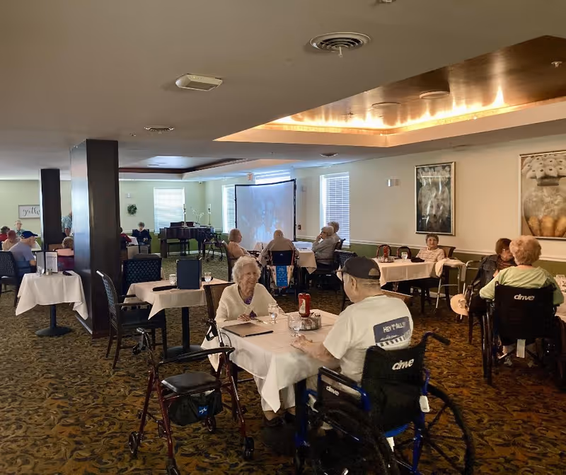 A dining room in a senior living facility with elderly residents seated at tables covered with white tablecloths. Some residents are in wheelchairs, and others use walkers. The room has carpeted floors, framed artwork on the walls, and a piano in the background. A projector screen is also visible at the far end of the room.