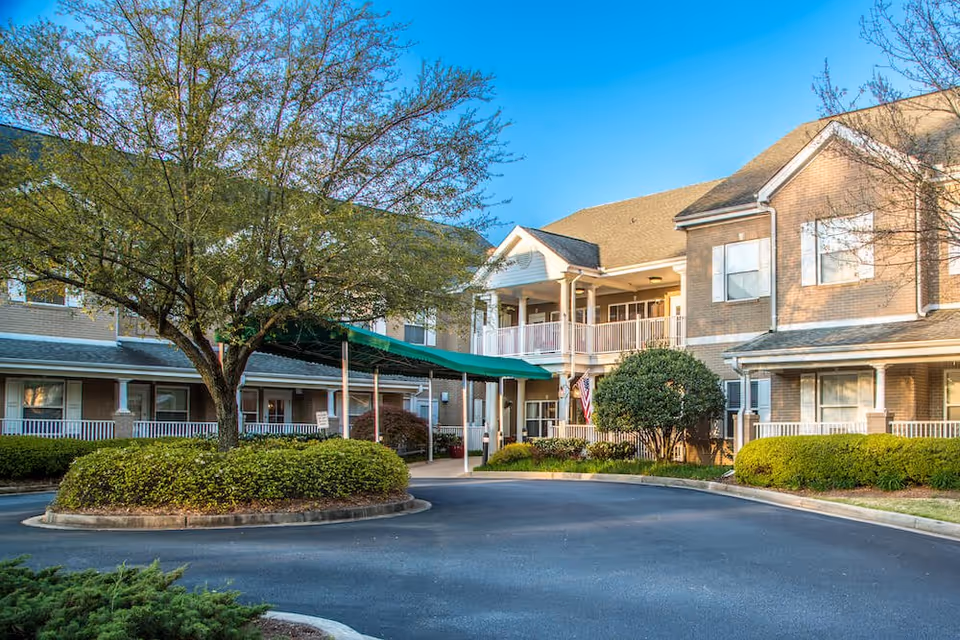 Exterior view of The Gardens at Eastside senior living facility showing a two-story brick building with white railings, a covered entrance with a green awning, landscaped bushes, and a tree in the circular driveway under a clear blue sky.