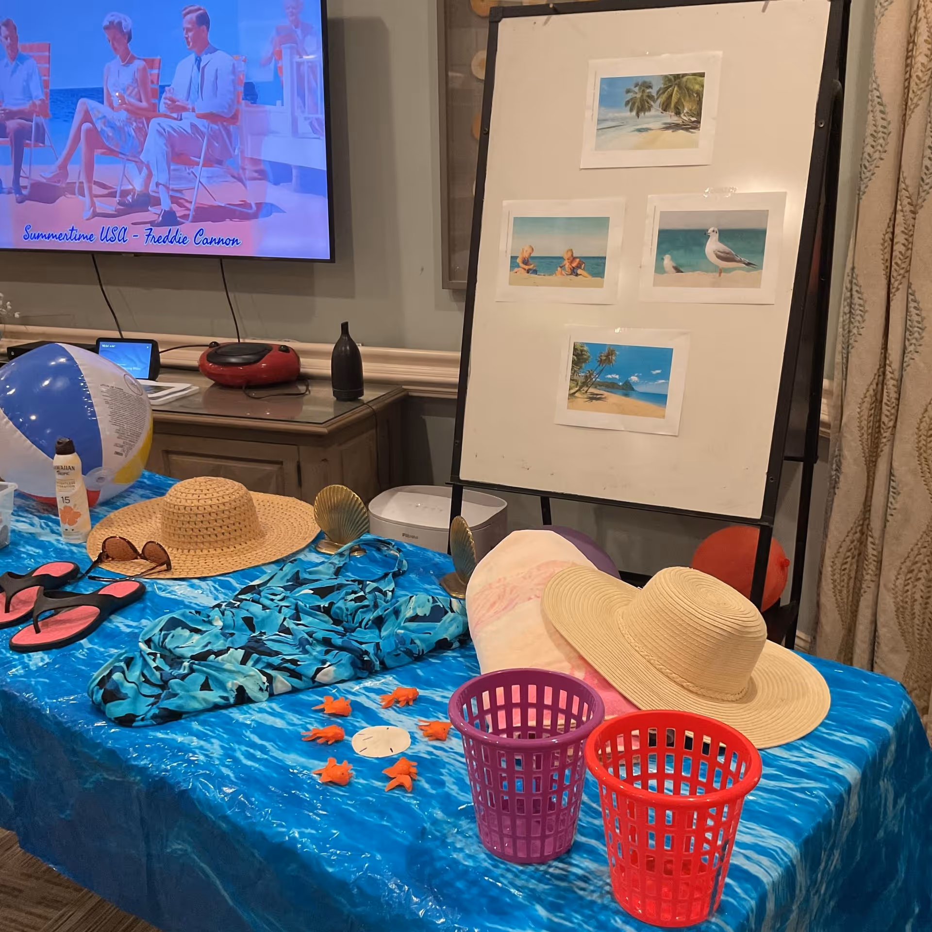 A table covered with a blue ocean-themed tablecloth displaying summer items including two straw hats, a pair of sunglasses, a beach ball, sunscreen, flip-flops, a blue patterned shirt, small orange starfish decorations, and two small plastic baskets. Behind the table is a whiteboard with four beach-themed photos attached. A TV screen on the wall shows an image of people sitting outdoors with the text 'Summertime USA - Freddie Cannon'.