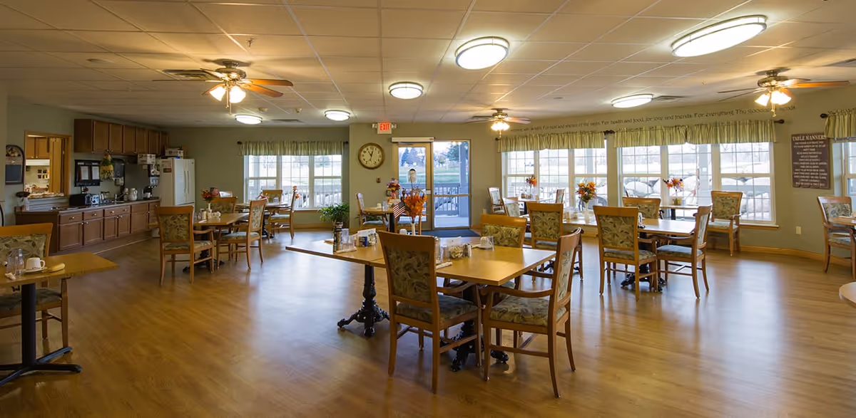 A spacious dining room in a senior living facility with multiple wooden tables and chairs arranged neatly. The room has large windows allowing natural light to fill the space, ceiling fans with lights, and a kitchen area with cabinets and appliances along one wall. The floor is wooden, and there are floral centerpieces on the tables.