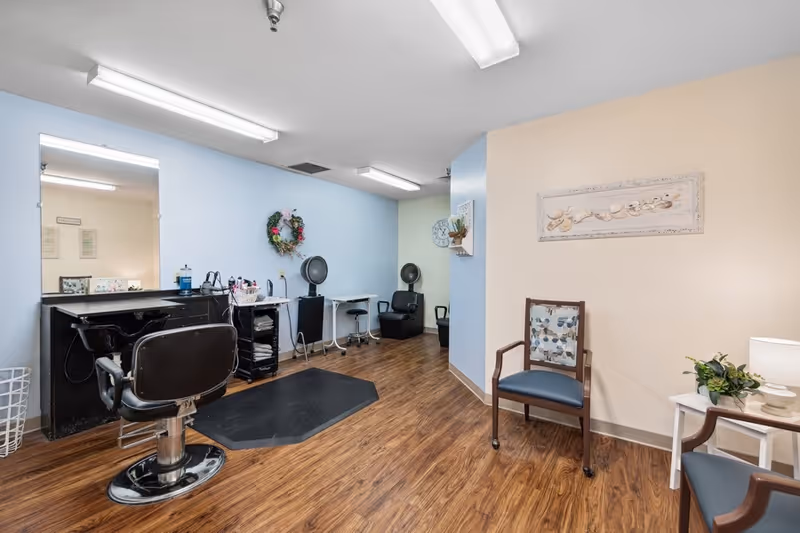 Interior view of a salon area in a senior living facility with a hair washing station, salon chairs, hair dryers, and seating area with chairs and a small table with a lamp and plant. The walls are painted light blue and beige, and the floor is wood.