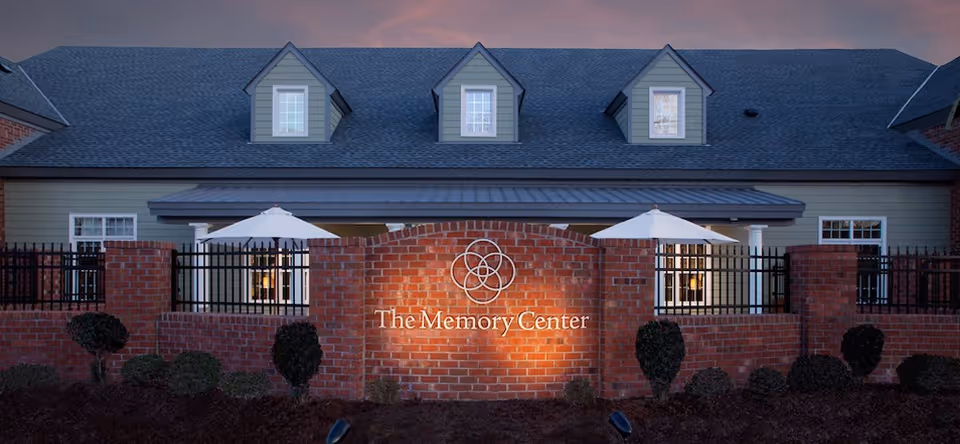 Exterior view of a brick building with a sign that reads 'The Memory Center' illuminated on a brick wall in front. The building has a gray roof with three dormer windows and two white umbrellas visible behind a black metal fence.