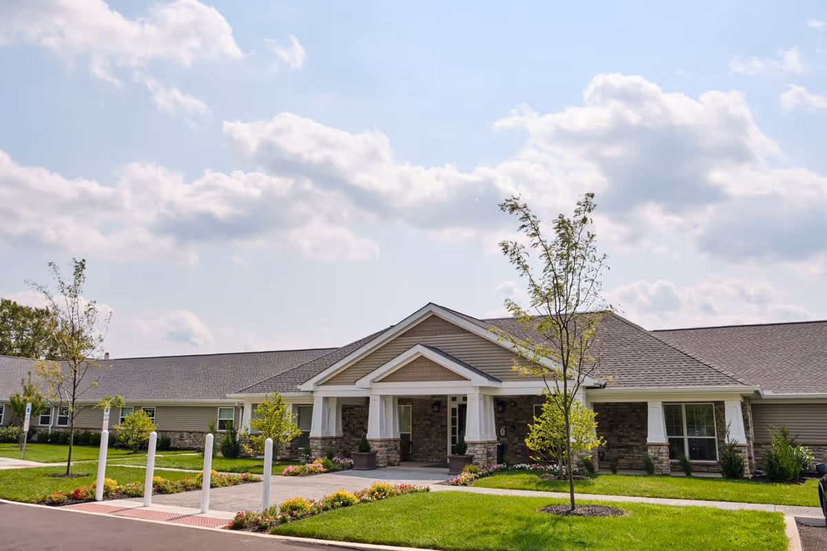 Exterior view of a single-story senior living facility building with a covered entrance, stone and siding facade, landscaped lawn with small trees and flower beds, under a partly cloudy sky.