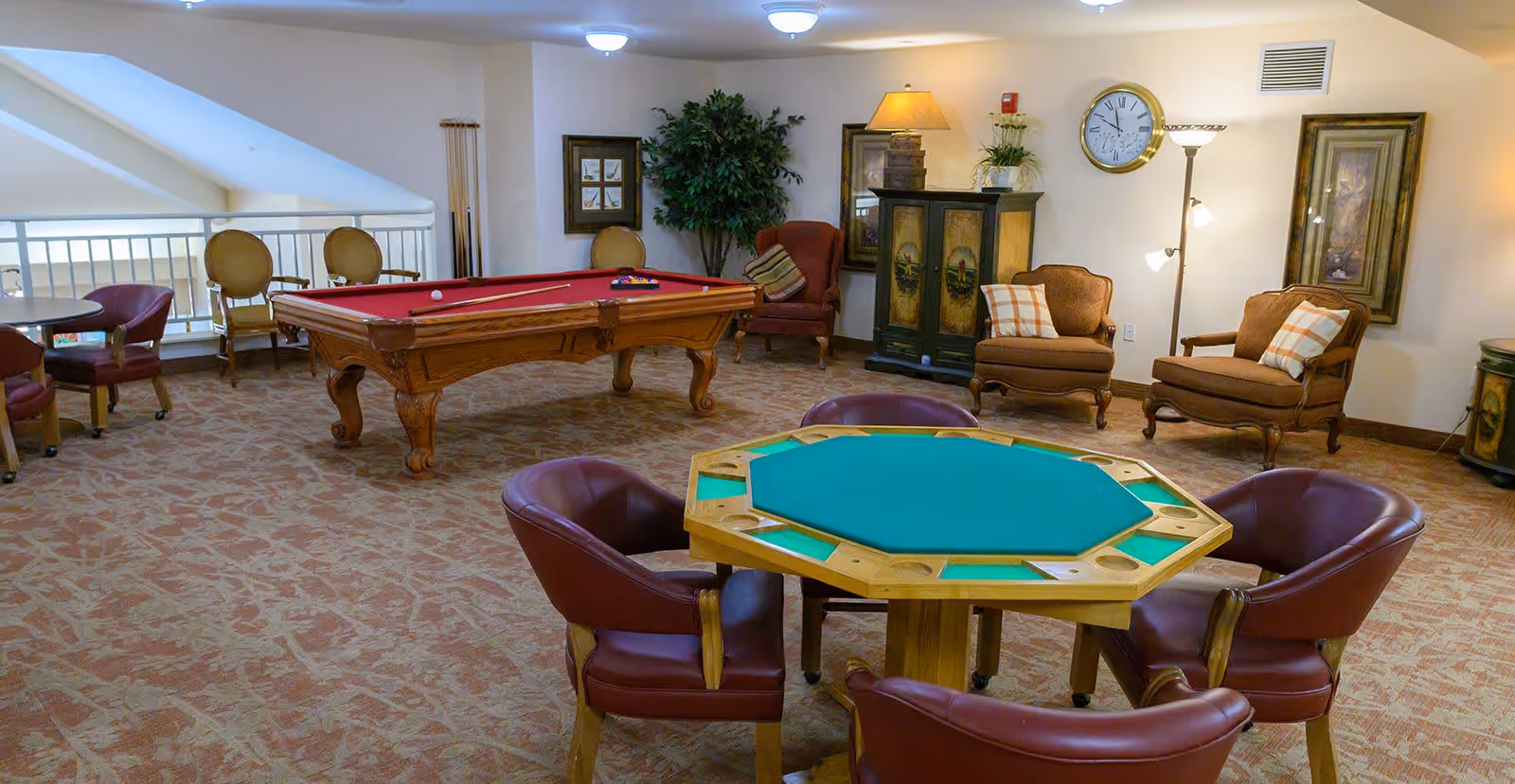 A recreational room with a red felt pool table, an octagonal card table with green felt and four leather chairs, several armchairs with cushions, framed artwork on the walls, a floor lamp, a clock, and a potted plant.