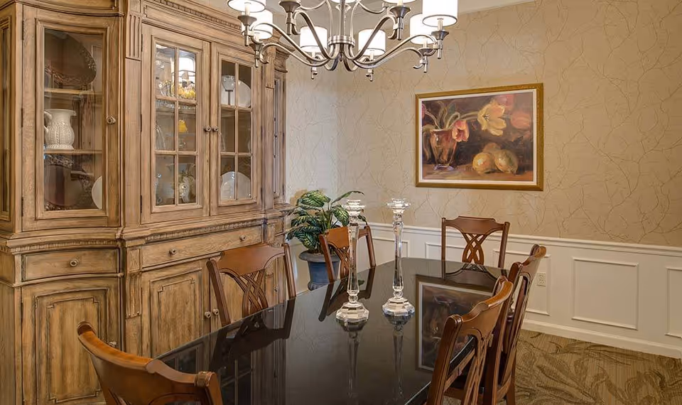 A dining room with a polished black dining table surrounded by wooden chairs. A wooden cabinet with glass doors displaying dishes and decorative items is against the wall. A chandelier with multiple lights hangs above the table. There is a framed painting of flowers and fruit on the wall, and a potted plant in the corner.