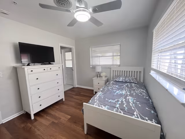 A small, bright bedroom with a single white bed featuring a gray and white patterned bedspread. There is a white nightstand with a lamp and decorative items next to the bed. Across from the bed is a white dresser with a flat-screen TV on top. The room has wooden flooring, white walls, a ceiling fan with a light, and two windows with blinds letting in natural light.