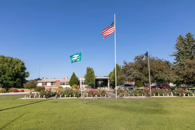 Exterior view of Parke View Rehabilitation & Care Center with a well-maintained lawn, flower beds, and three flagpoles displaying the American flag, a green and white flag, and a blue flag. The building is single-story with trees and parked cars visible in the background under a clear blue sky.