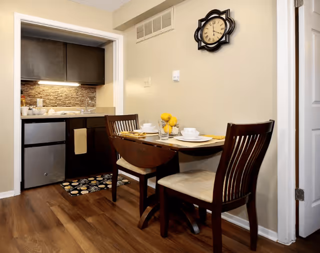 A small dining area with a wooden table set for two, featuring white plates, bowls, and cups, along with yellow napkins and a centerpiece of yellow flowers. The dining area is adjacent to a compact kitchen with dark cabinets, a small refrigerator, a sink, and a stone tile backsplash. A decorative wall clock is mounted above the dining table on a beige wall.
