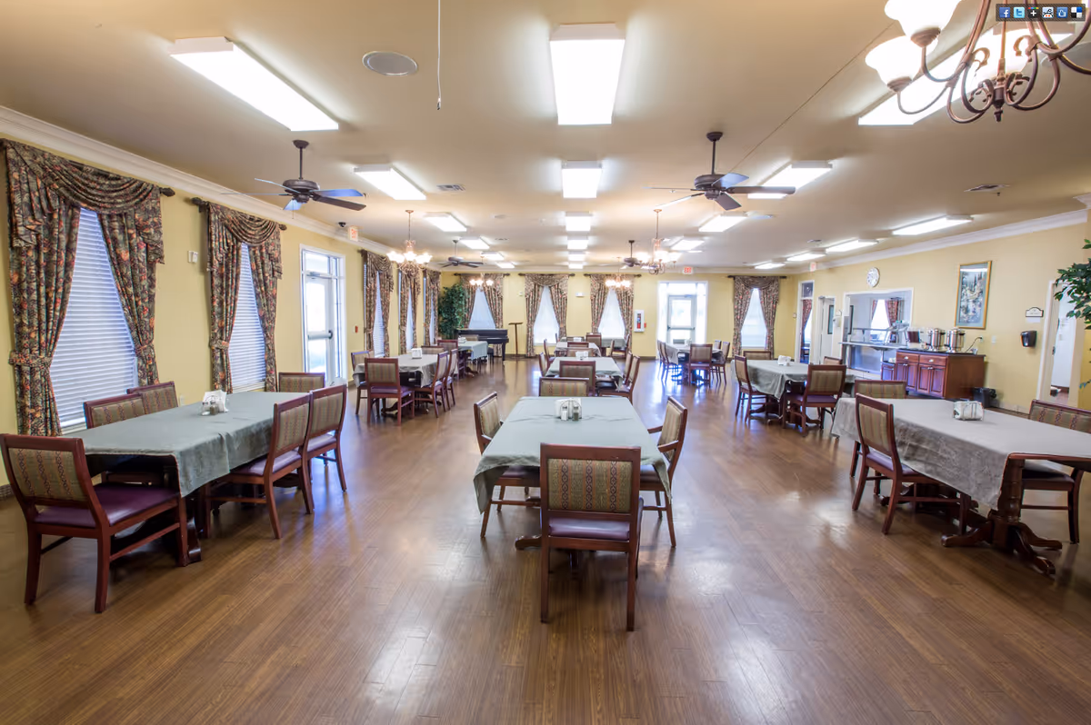 A spacious dining room with multiple rectangular tables covered with green tablecloths and surrounded by wooden chairs with patterned upholstery. The room has large windows with floral curtains, ceiling fans, chandeliers, and bright overhead lighting. There is a piano in the back corner and a serving area with food warmers on the right side.