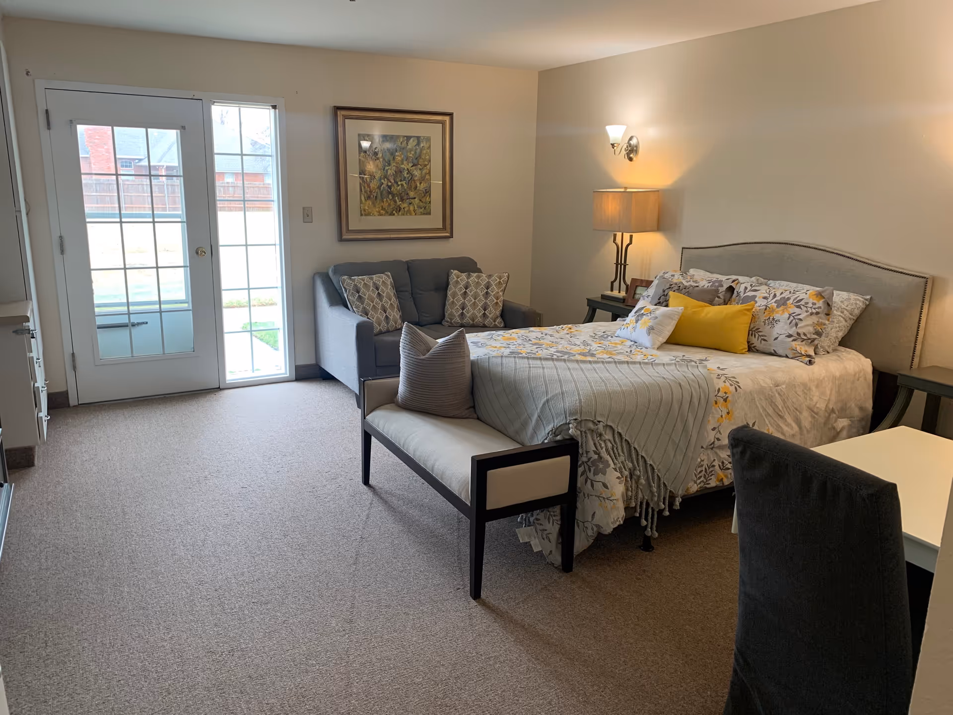 A cozy bedroom in an assisted living facility featuring a bed with floral bedding and multiple pillows, a small bench at the foot of the bed, a gray loveseat with patterned cushions, a framed painting on the wall, a lamp on a side table, and a door with glass panes leading outside.