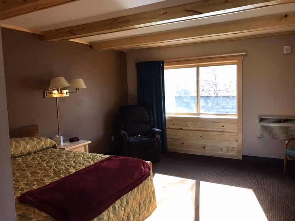 A cozy bedroom with a bed covered in a patterned yellow and green bedspread and a burgundy blanket at the foot. There is a wooden nightstand with a lamp and a small item on it. A dark recliner chair is positioned near a window with dark curtains, allowing sunlight to brighten the room. The walls are painted brown and the ceiling has exposed wooden beams.