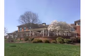 Exterior view of a brick building with a covered patio area surrounded by shrubs and a blooming tree, under a clear sky.