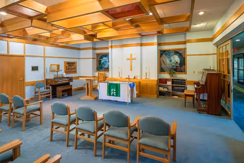 Interior view of a small chapel or worship room with wooden ceiling beams, a cross on the wall behind an altar covered with a white cloth and green cloth with religious symbols. There are several rows of wooden chairs with blue cushions facing the altar. The room also contains two pianos, stained glass windows, and shelves with books and hymnals.