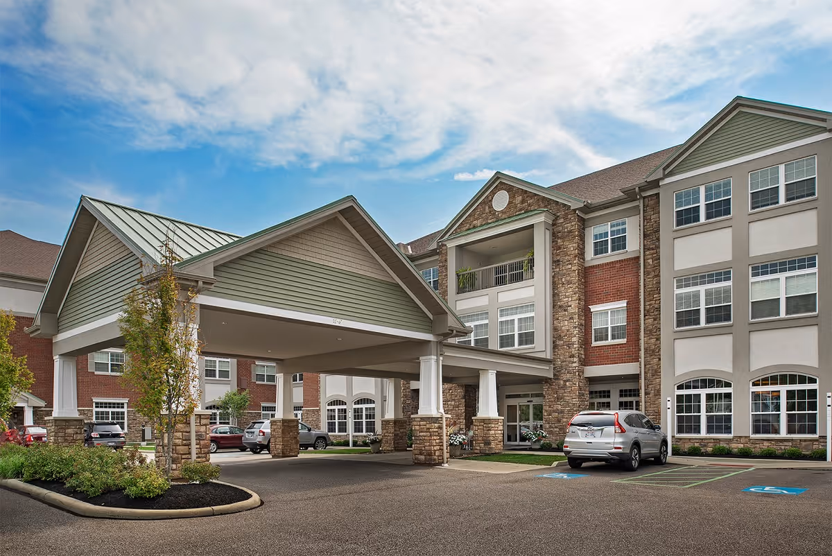 Exterior view of a senior living facility named Summit Corners showing a covered entrance with stone pillars, multiple windows, and parked cars in front under a partly cloudy sky.