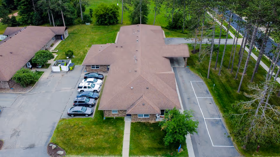 Aerial view of a senior living facility building with a brown roof, surrounded by green lawns and trees. Several cars are parked in a parking lot adjacent to the building. A sidewalk leads to the entrance of the building.