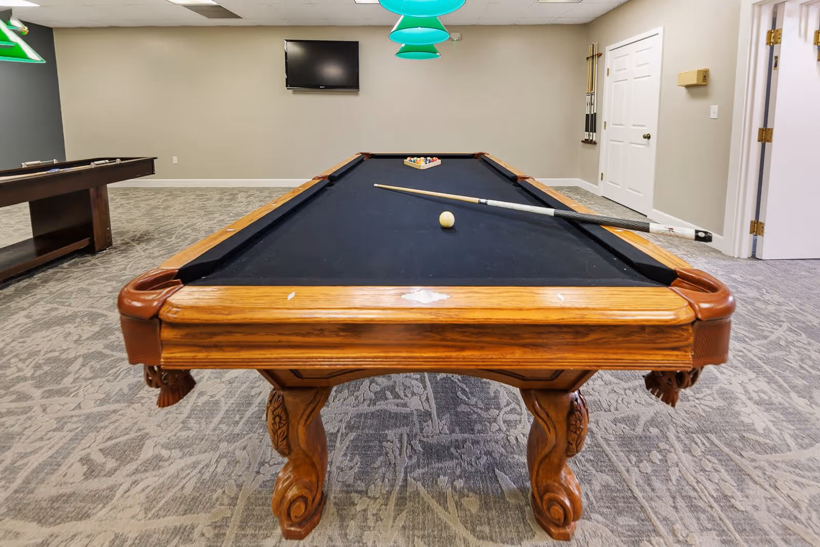 A recreational room with a wooden pool table featuring a black felt surface. A cue stick and a white cue ball are on the table, with a rack of billiard balls at the far end. The room has carpeted flooring, beige walls, a wall-mounted TV, and a set of pool cues hanging on the wall. Green hanging lights are visible above the pool table.