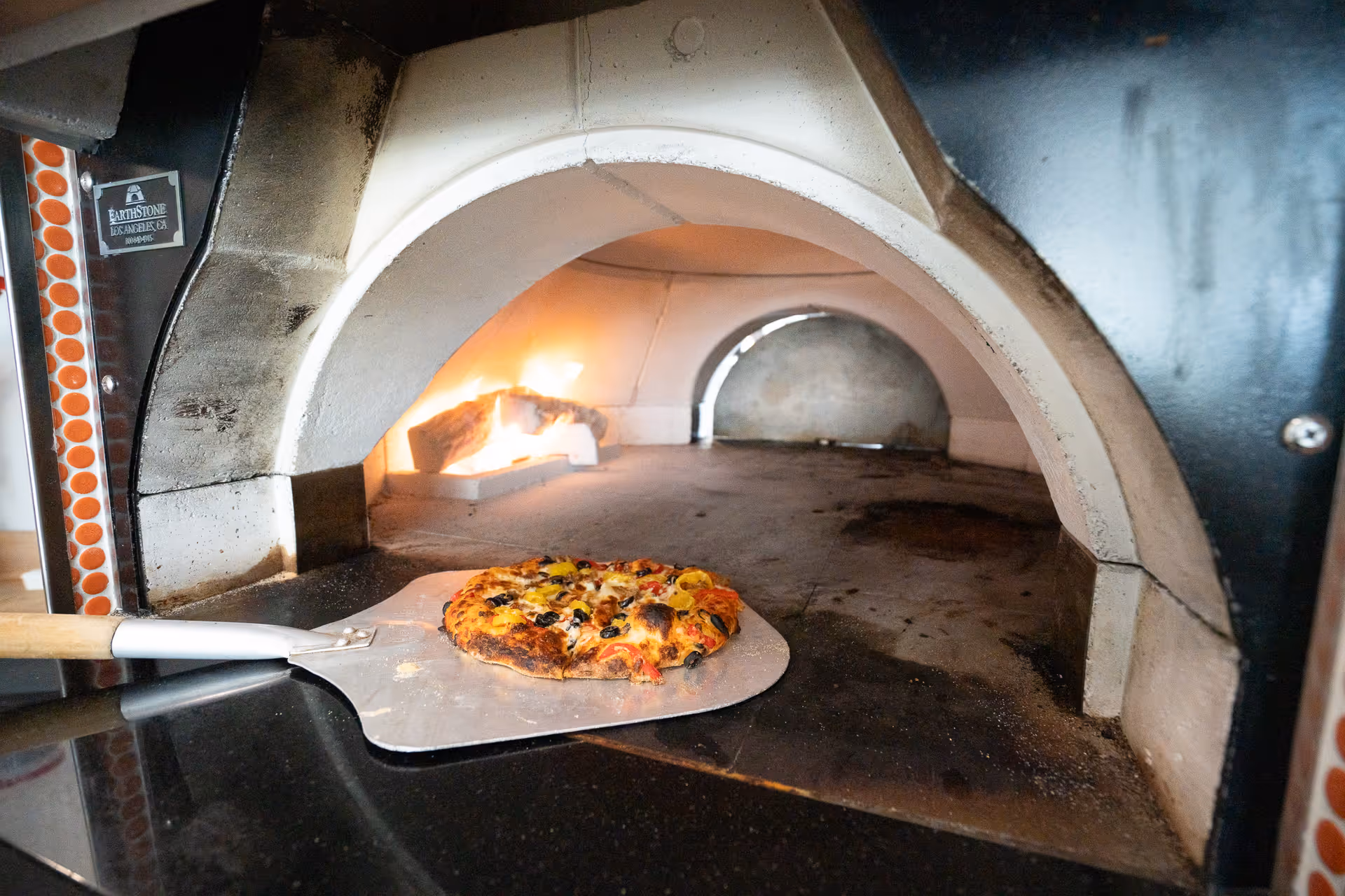 A wood-fired pizza oven with a pizza peel holding a freshly baked pizza inside the oven. The oven has a visible fire burning in the back.