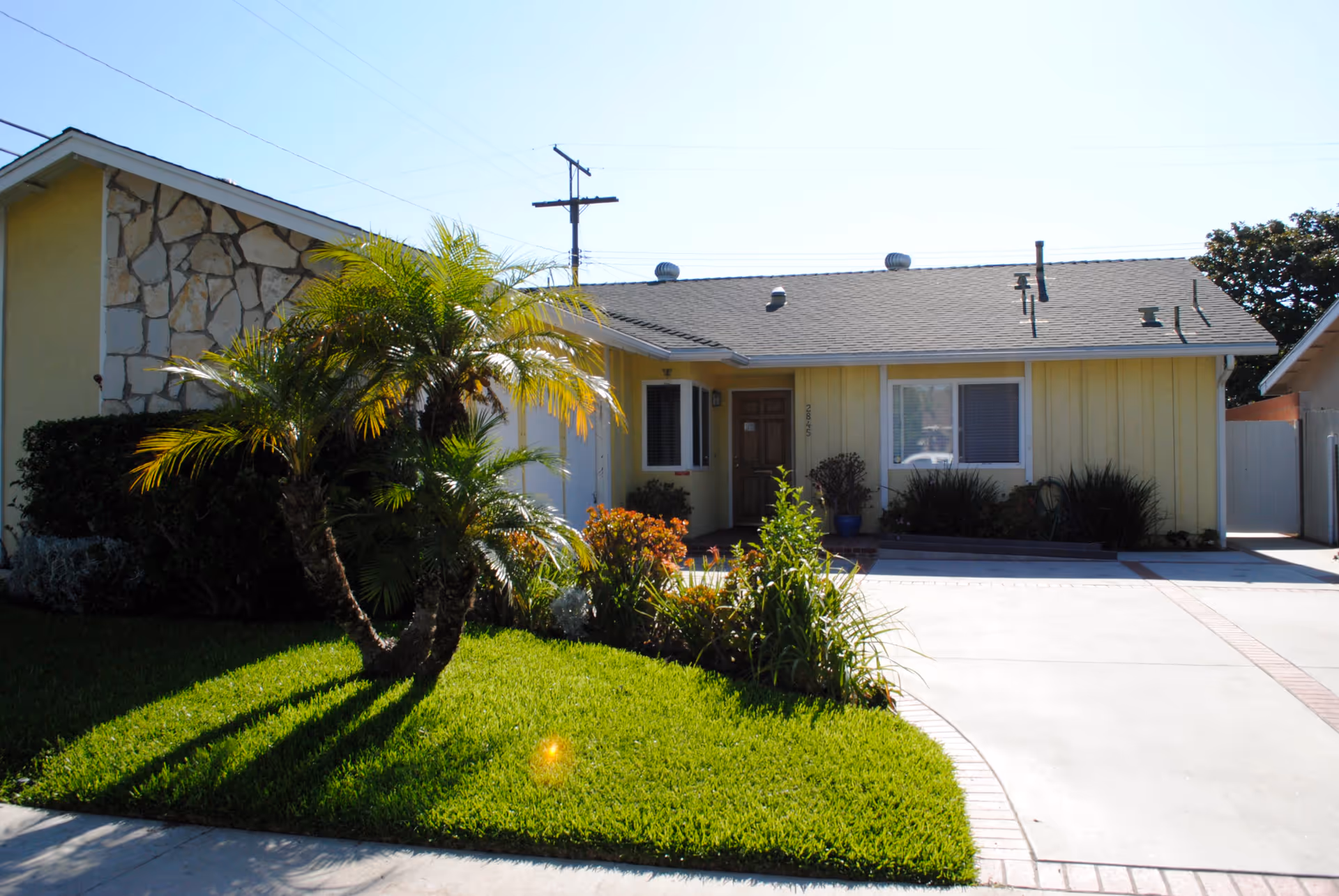 Front exterior view of a single-story yellow house with a stone accent wall on the left side, a brown front door, a window with blinds, and a well-maintained lawn with palm trees and shrubs. The driveway is concrete with brick trim.