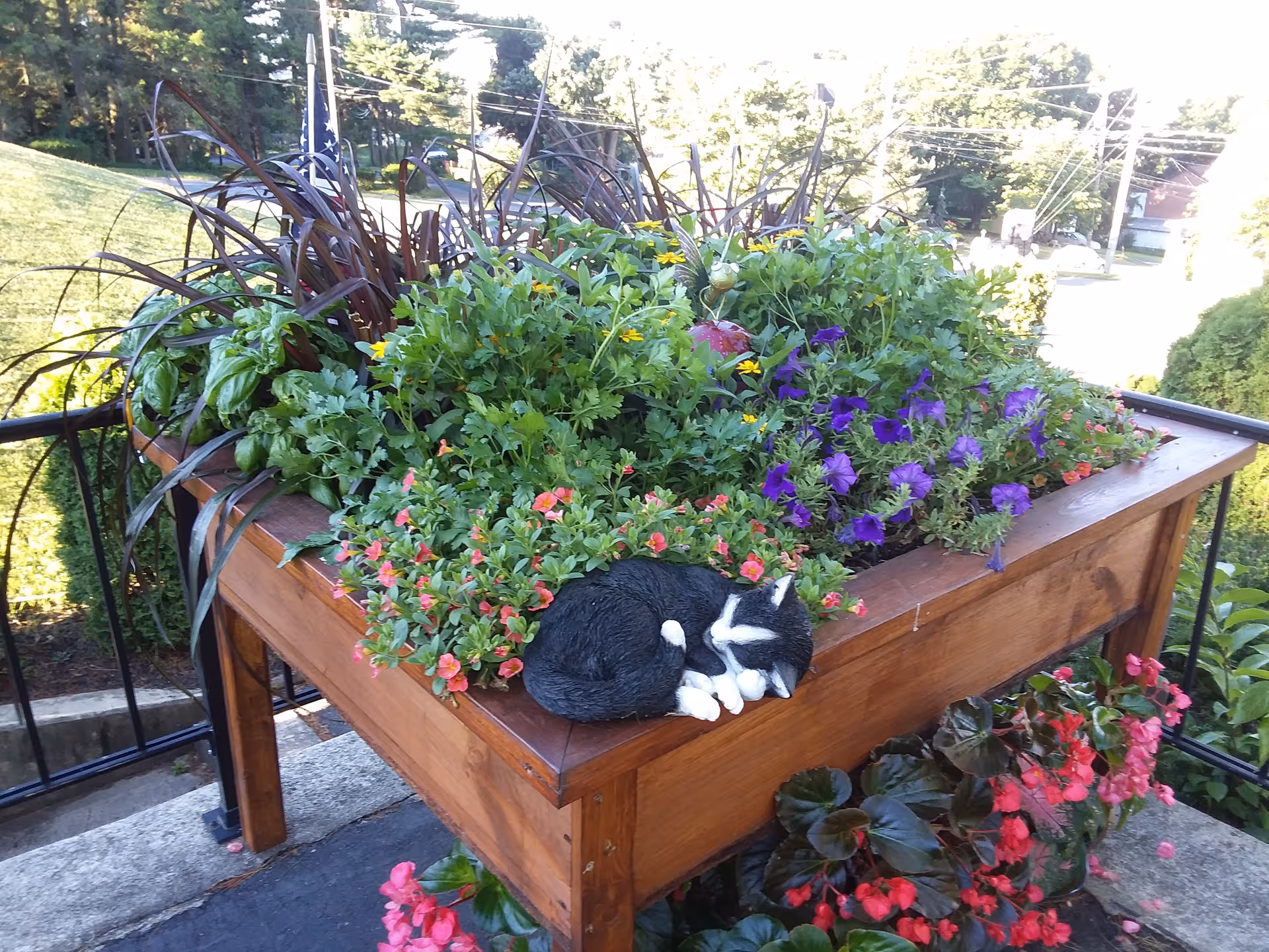 Wooden raised planter on a porch overflowing with colorful flowers and a small sleeping cat statue on its edge.