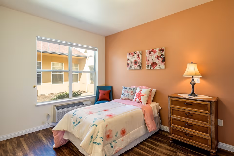 A cozy bedroom with a single bed adorned with floral bedding and decorative pillows, including one with a starfish design. The room has a wooden floor, a wooden dresser with a lamp on it, and two floral paintings on a warm orange accent wall. A large window lets in natural light and shows an exterior view of the building.