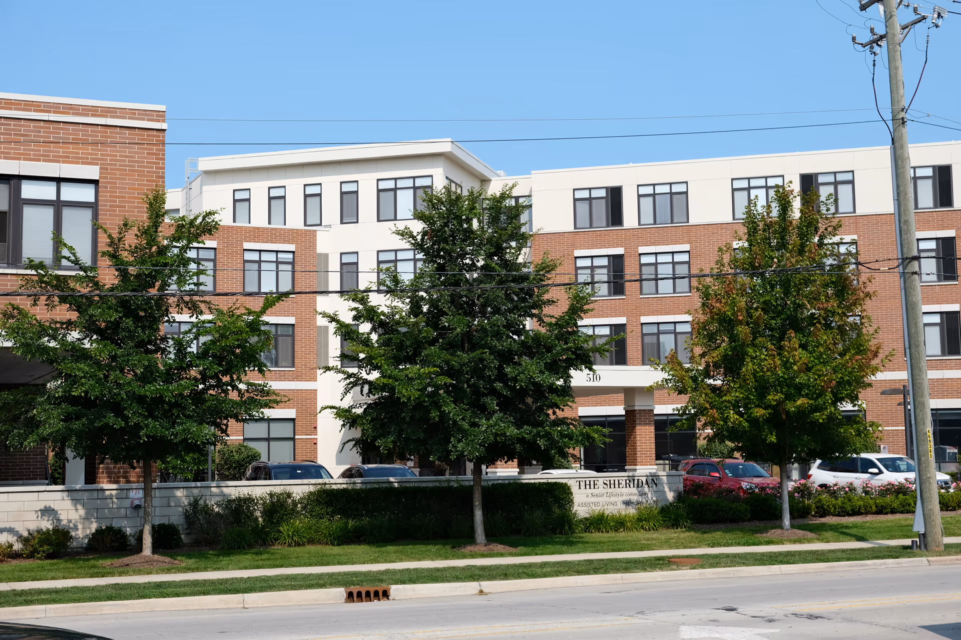 Front exterior of The Sheridan at Park Ridge multi-story brick and white senior living building with trees and parked cars.