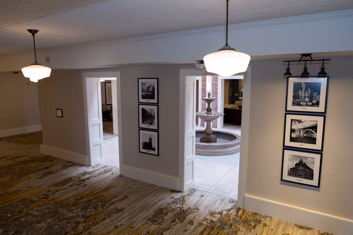 Interior hallway of Knoxville High Apartments with carpeted floor, two hanging ceiling lights, and two doorways leading to a courtyard area with a stone fountain. Black and white framed photographs are displayed on the walls beside the doorways.