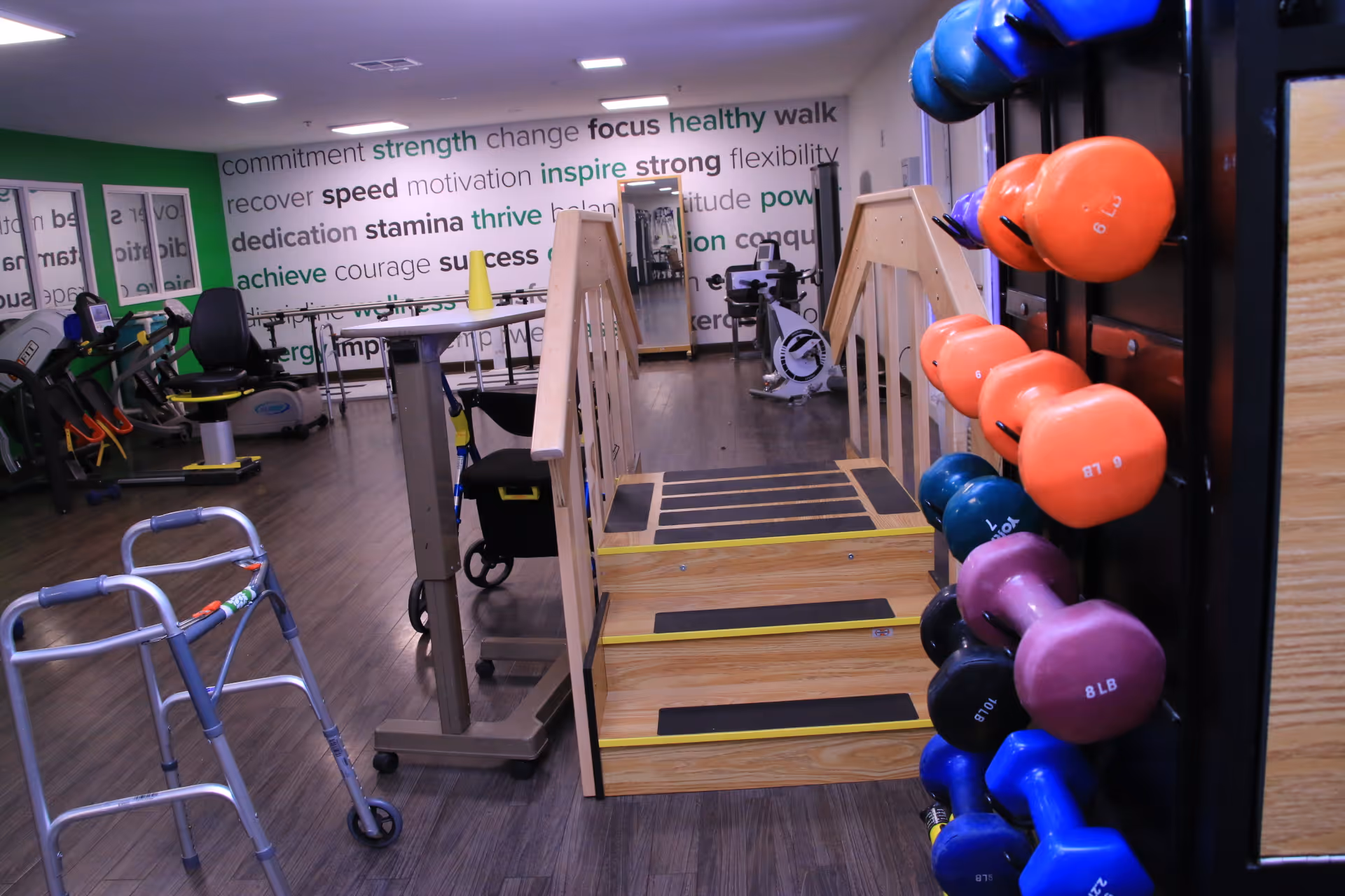Interior view of a physical therapy or fitness room in a senior living facility. The room features a wooden staircase with handrails for rehabilitation exercises, a walker, various exercise equipment including stationary bikes and parallel bars, and a rack of colorful dumbbells on the right side. The back wall has motivational words like strength, focus, inspire, and success displayed in various fonts and colors.
