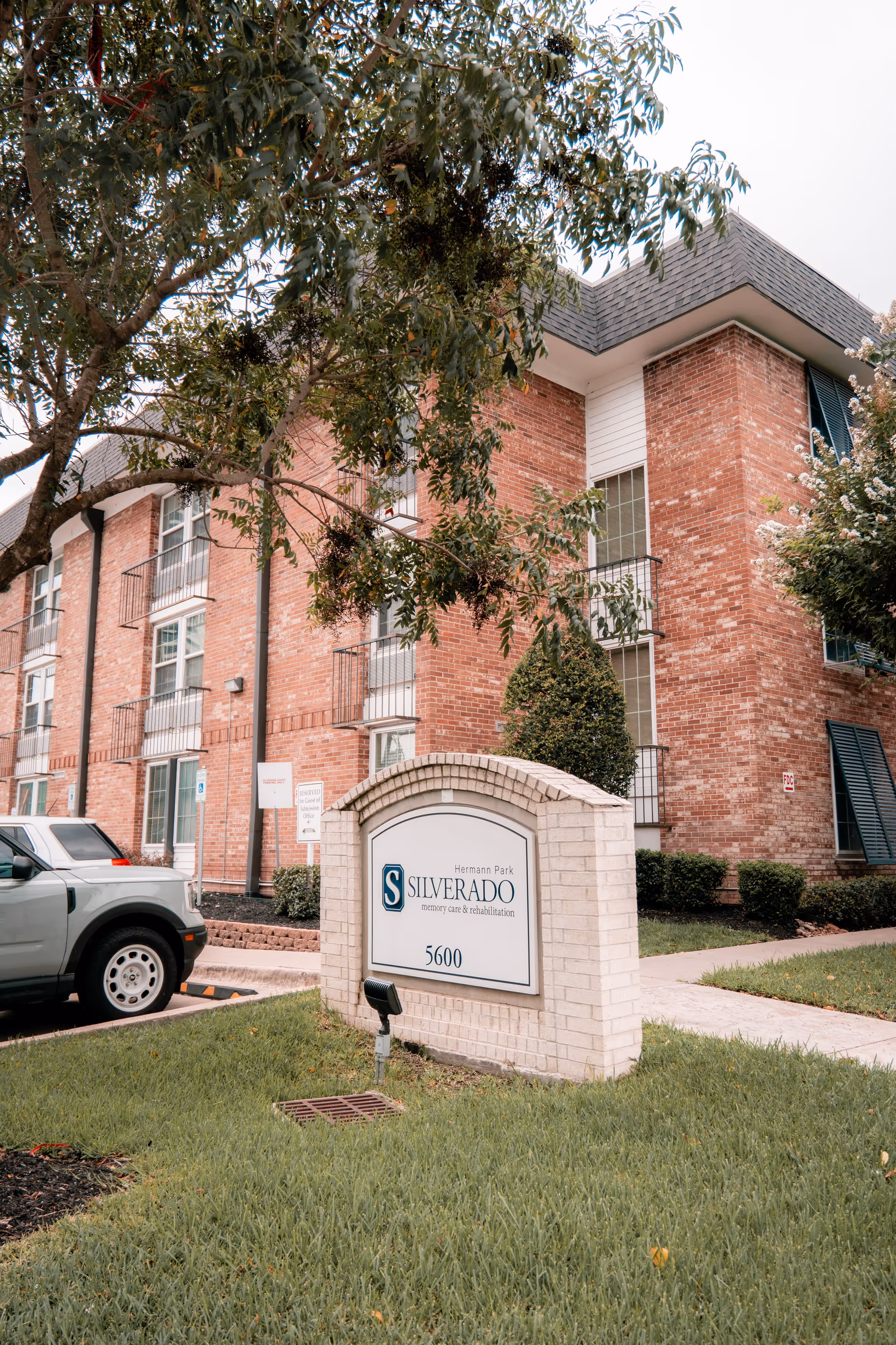 Exterior view of Silverado Hermann Park Memory Care Community building with a brick facade, multiple windows, and small balconies. In front of the building is a white brick sign displaying the facility name and address number 5600. There is a tree partially covering the view and a parked silver SUV on the left side.