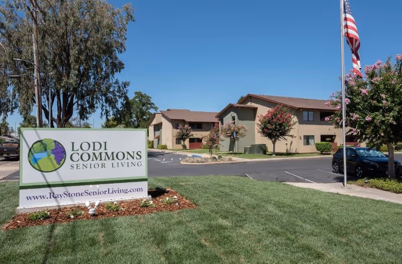Exterior view of Lodi Commons Senior Living facility with a large sign in the foreground displaying the facility name and website. The building is two stories with beige and green walls, surrounded by trees and a parking lot. An American flag is visible on a flagpole to the right.