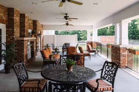 Covered outdoor patio area with brick pillars and walls, featuring multiple seating arrangements including cushioned chairs and metal tables with decorative patterns. Ceiling fans are installed overhead, and potted plants are placed on tables and along the edges. The area overlooks greenery beyond the railing.