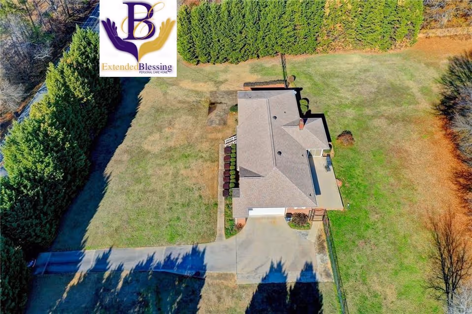 Aerial view of a single-story house surrounded by a large grassy yard with trees and bushes along the perimeter. The driveway leads to a garage attached to the house. The image includes the logo of Extended Blessing Personal Care Home in the top left corner.