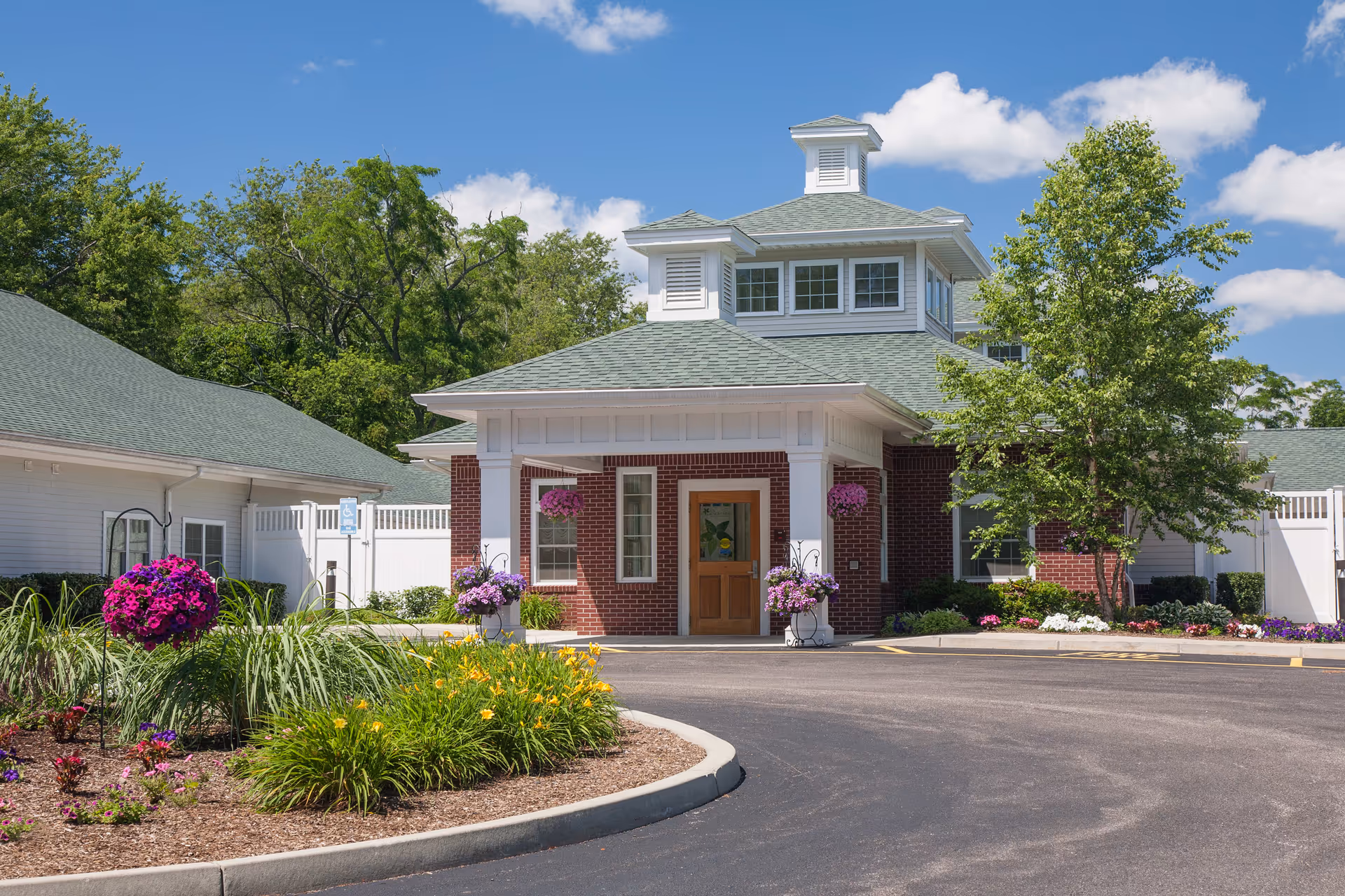 Front exterior view of a senior living facility building with a covered entrance, brick facade, and white trim. The driveway curves in front of the entrance, and there are landscaped flower beds with colorful flowers and greenery. Trees and a clear blue sky are visible in the background.