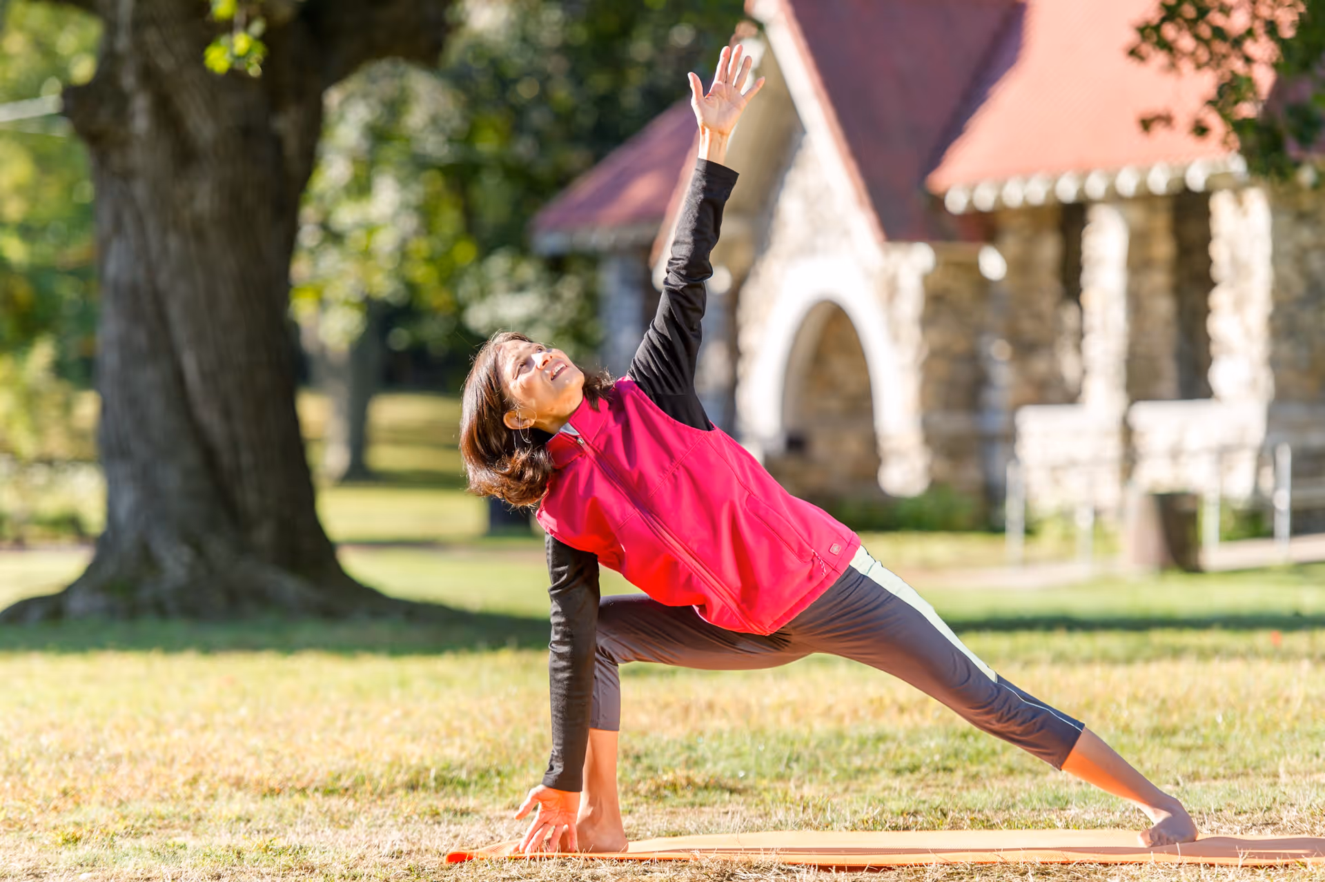 A woman wearing a red vest and gray pants is performing a yoga stretch on a mat outdoors in a grassy area with trees and a stone building with a red roof in the background.