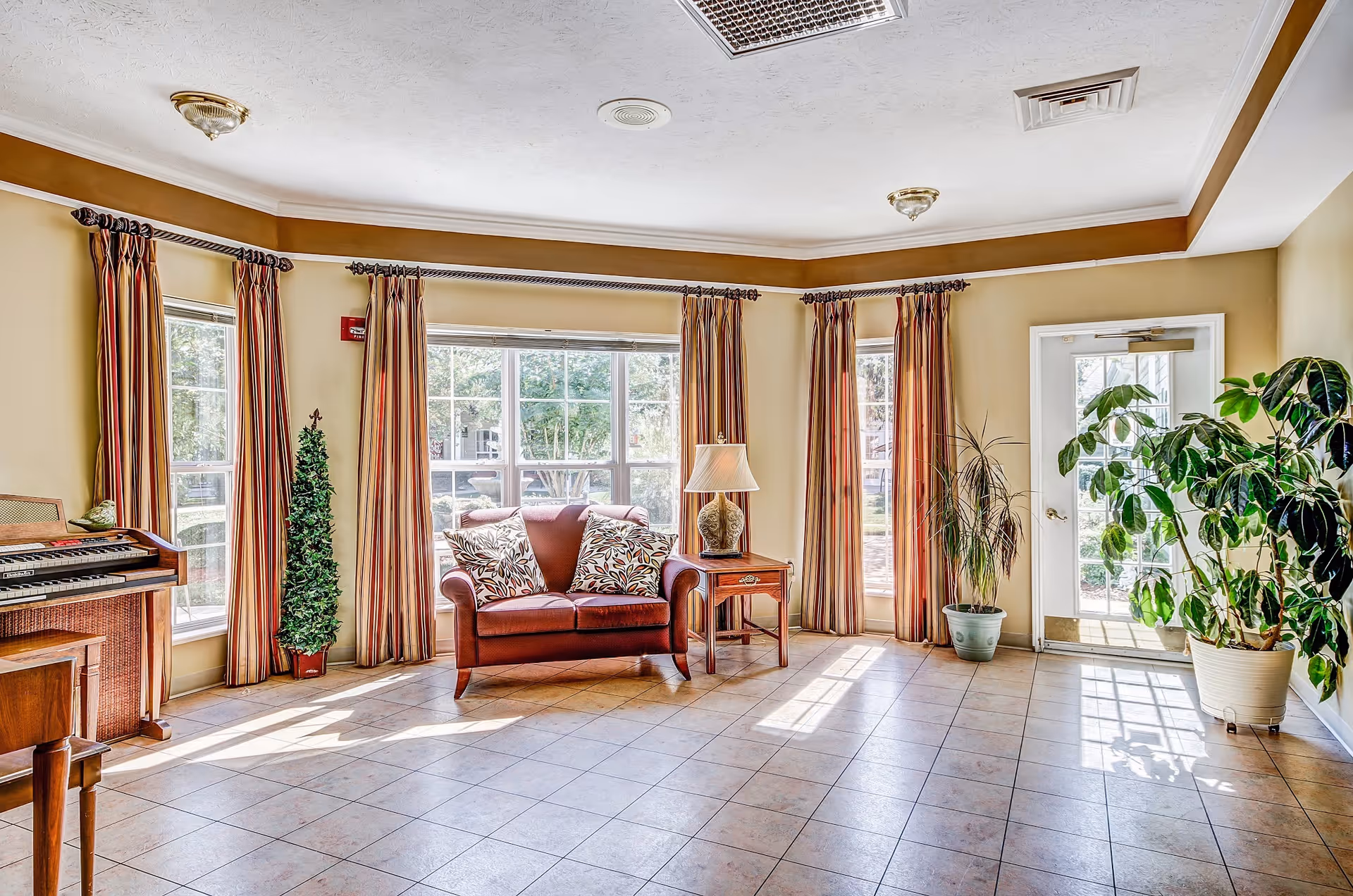 A bright and spacious living room area with large windows covered by striped curtains allowing natural light to fill the space. The room features a red loveseat with two patterned pillows, a wooden side table with a decorative lamp, several potted plants, and a wooden organ on the left side. The floor is tiled, and there is a glass door leading outside.