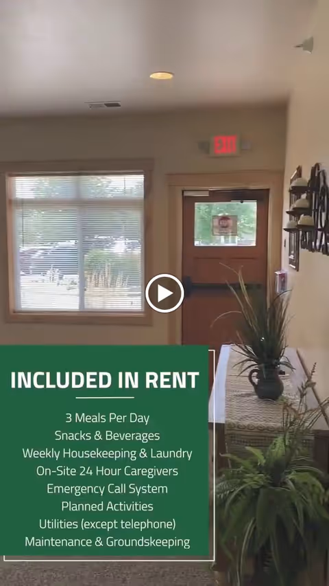 Interior view of an assisted living facility entrance area showing a wooden door with a window, a window with blinds, a table with a plant on it, and wall-mounted candle holders. A green text box lists amenities included in rent such as meals, housekeeping, caregivers, and maintenance.