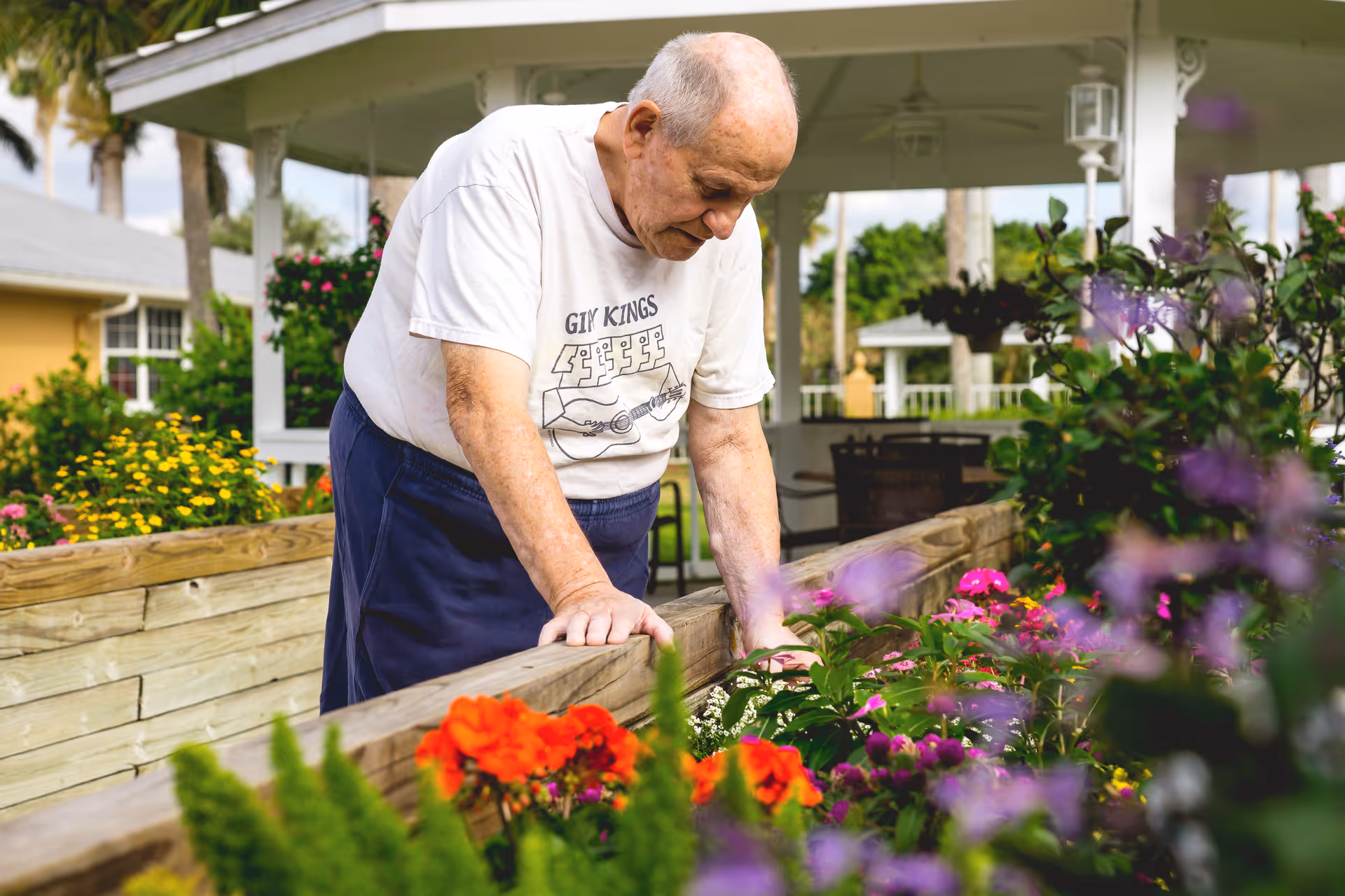 An elderly man wearing a white t-shirt and dark pants is tending to a raised garden bed filled with colorful flowers. The setting is outdoors with a covered patio and greenery in the background.