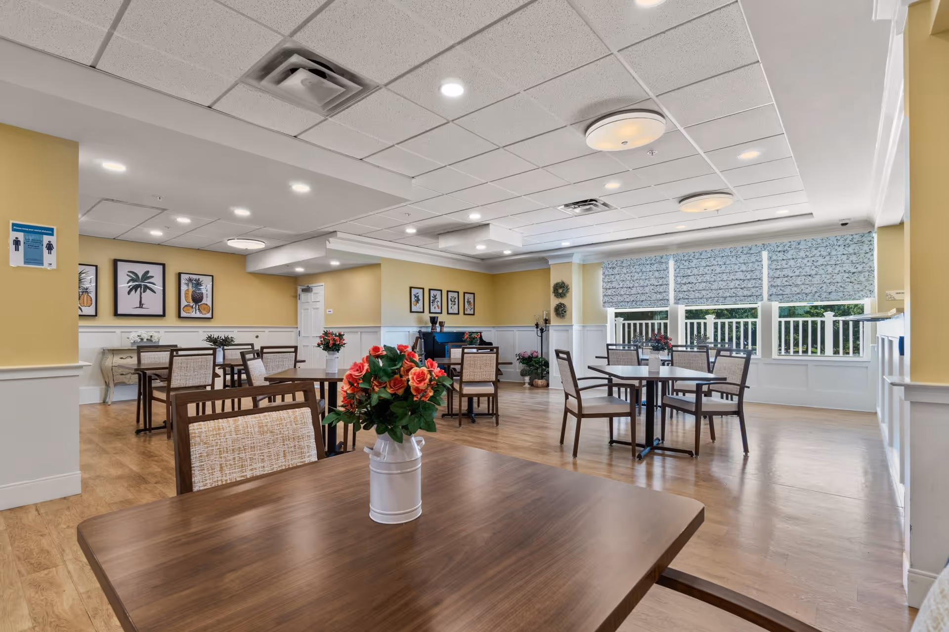 A bright and spacious dining room in a senior living facility with multiple wooden tables and chairs. Each table has a vase with red and pink flowers. The walls are painted yellow with white wainscoting, and framed botanical artwork decorates the walls. Large windows with patterned blinds allow natural light to fill the room. The ceiling has recessed lighting and modern light fixtures.