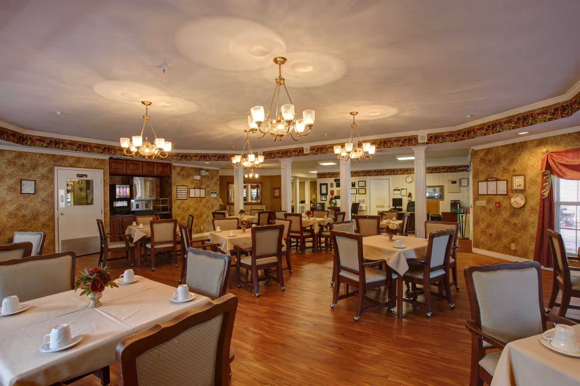 Well-lit dining room with multiple tables and chairs, chandeliers, and place settings in a senior living facility.