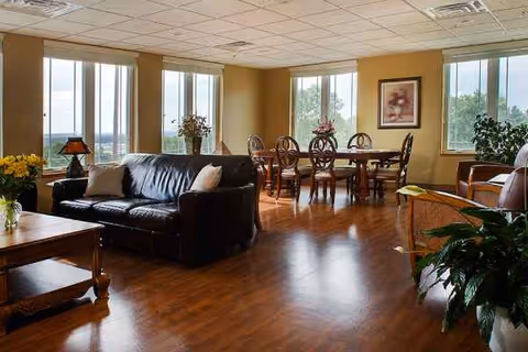 Spacious senior living lounge with leather sofa, wooden tables, plants, and large windows.