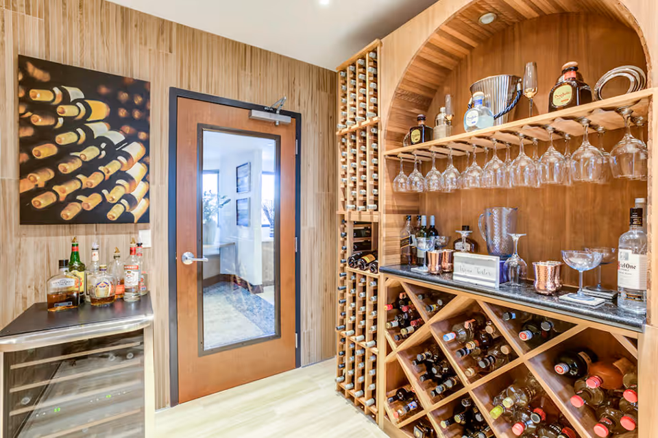 A well-organized wine storage room with wooden racks filled with wine bottles, a countertop holding various liquor bottles and glassware, and a glass-paneled wooden door leading to another room. The walls have a wood panel design and there is a painting of wine bottles on the left wall.