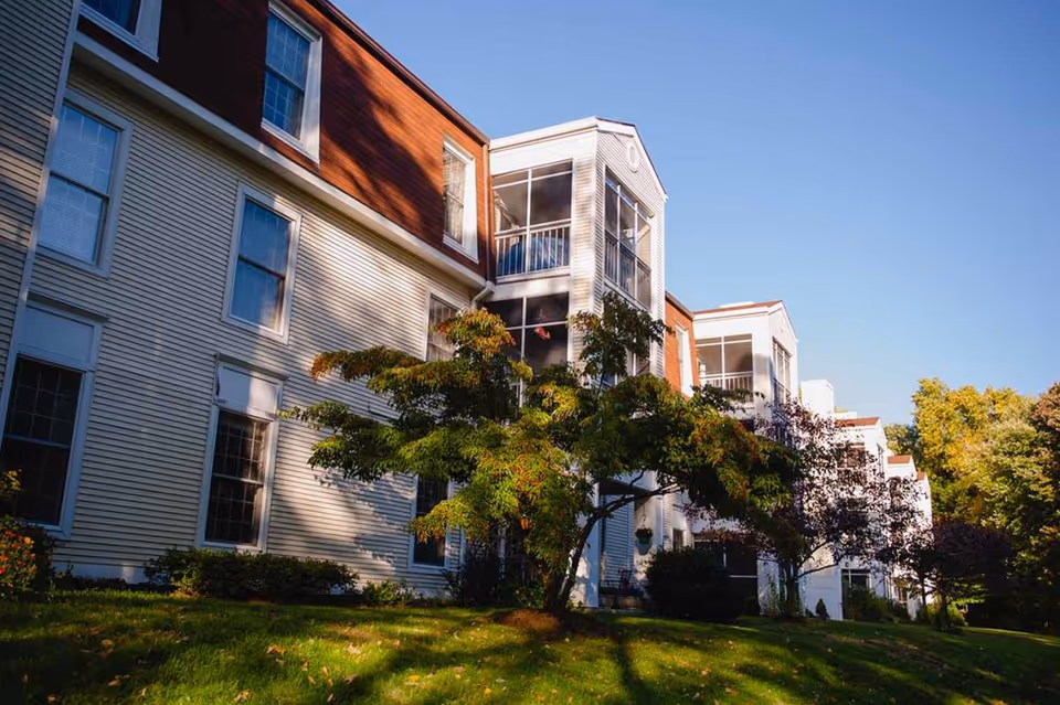 Exterior view of a multi-story residential building with white siding and red-brown accents, surrounded by green grass and trees under a clear blue sky.