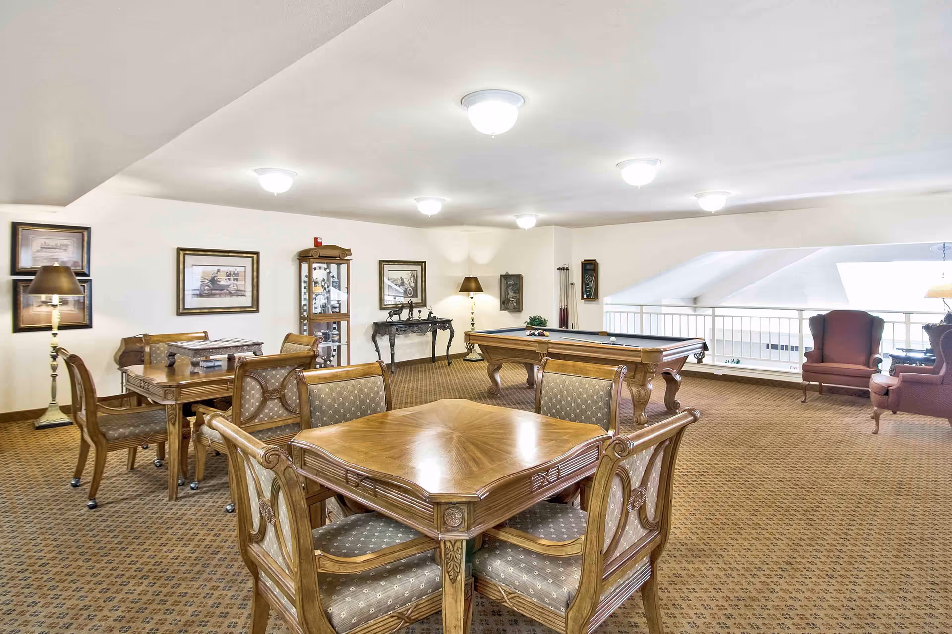 Bright communal activity room with wooden card tables and chairs, a pool table, and lounge seating.