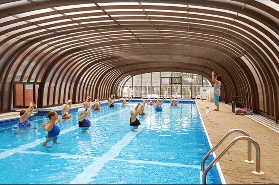 A group of elderly women participating in a water exercise class in an indoor swimming pool with a curved, transparent roof. An instructor stands on the pool deck guiding the class.