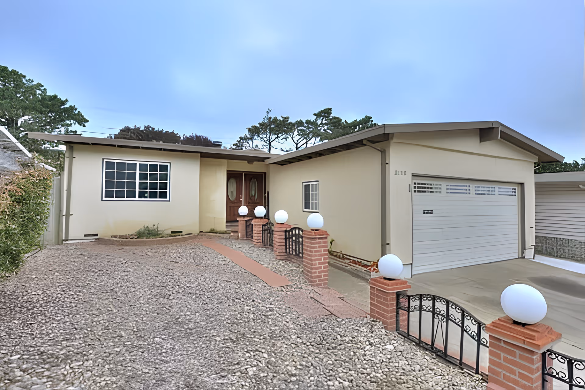 Exterior view of a single-story residential building with a beige facade, a double garage door, a large window, and a front entrance with double wooden doors. The driveway is gravel with a paved walkway leading to the entrance, bordered by brick pillars topped with round white lights and black metal gates. Trees are visible in the background under a clear blue sky.