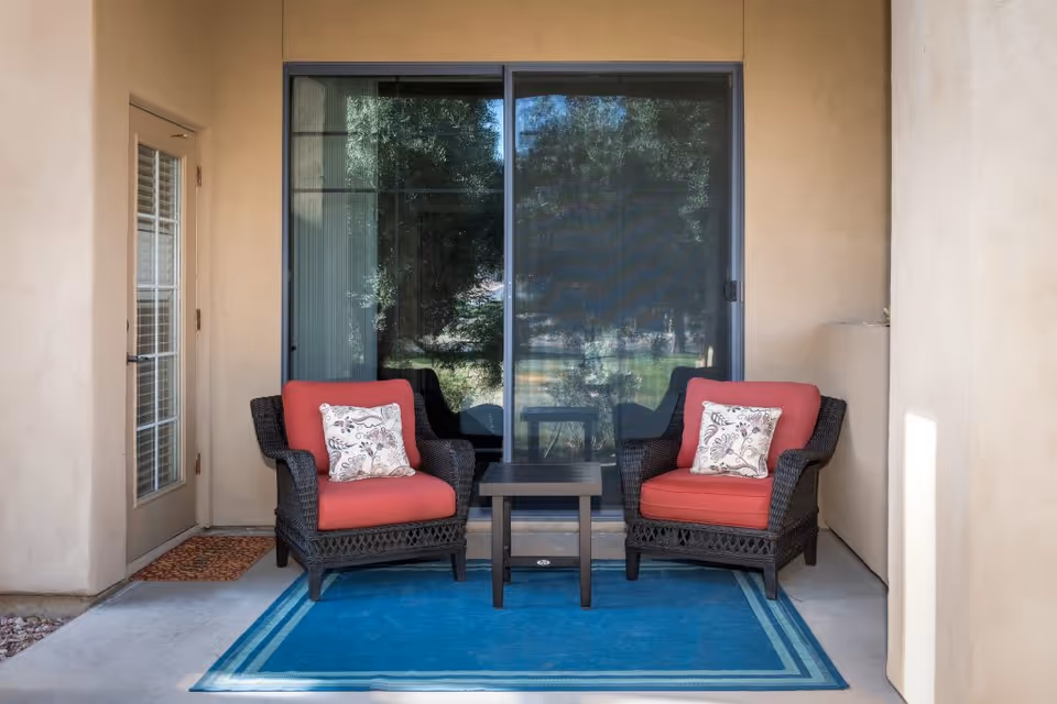 A small outdoor patio area with two dark wicker chairs featuring red cushions and floral patterned throw pillows, separated by a small dark wooden table. The patio has a blue rug with a double border design, a glass sliding door behind the chairs, and a beige stucco wall with a door on the left side.