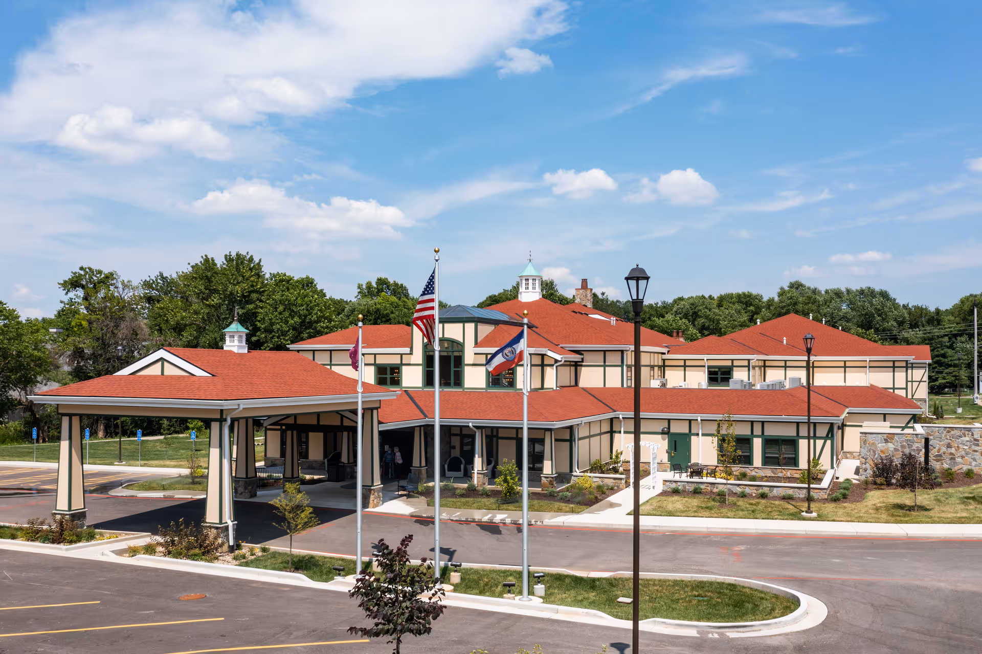 Exterior view of Creekside at Elfindale Assisted Living facility showing a large building with red roofs, beige walls with green trim, a covered entrance, three flagpoles with flags, and a parking area under a partly cloudy blue sky.