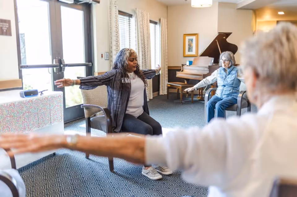 A group of elderly women seated in chairs in a well-lit room participating in a seated exercise or stretching activity led by an instructor. The room has large windows with curtains, a piano in the background, and a table with a colorful tablecloth near the door.