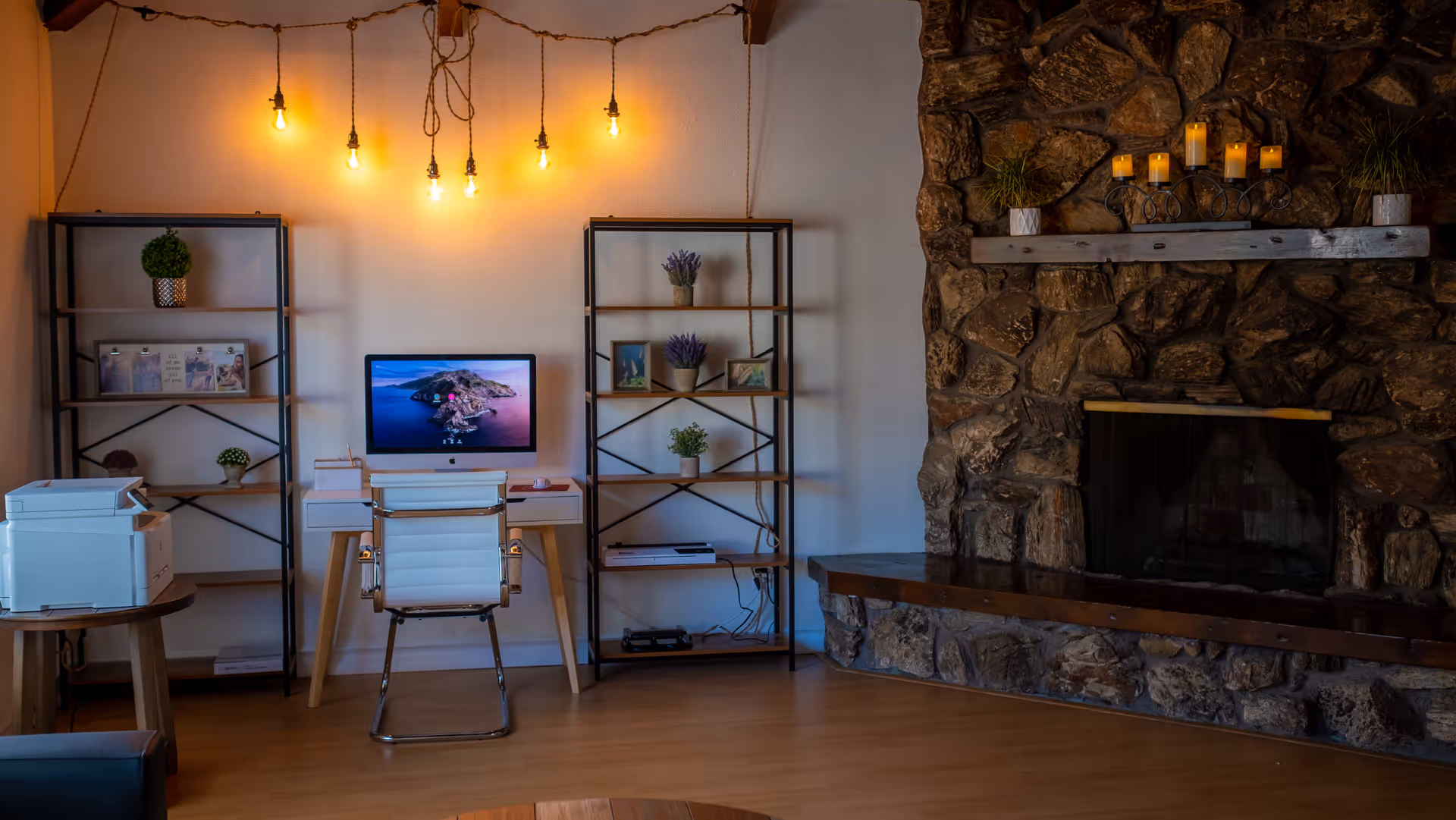A cozy interior room featuring a white desk with a computer and a white chair in front of it. On either side of the desk are black metal and wood shelves decorated with small plants, framed photos, and books. Above the desk, several hanging light bulbs provide warm lighting. To the right, there is a large stone fireplace with a wooden mantel adorned with candles and small potted plants. The floor is wooden, and a printer sits on a small round wooden table to the left.