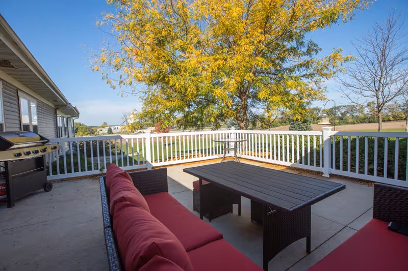Outdoor patio area with a red cushioned wicker sofa and matching chairs around a rectangular table. There is a white railing enclosing the patio, a barbecue grill on the left side, and a large tree with yellow leaves in the background under a clear blue sky.